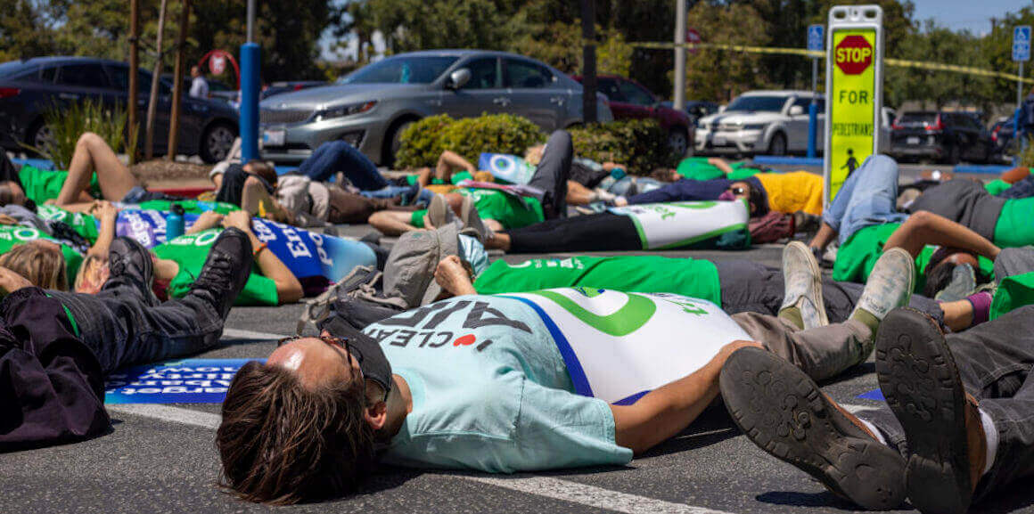 Several protesters lie face up on a Target parking lot pavement in Long Beach, California. At the center, there is a man wearing a black mask over his mouth and nose and a light blue t-shirt with a campaign poster placed on top of his body.
