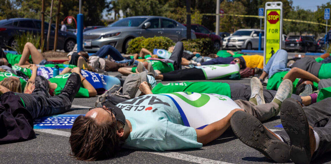 Several protesters lie face up on a Target parking lot pavement in Long Beach, California. At the center, there is a man wearing a black mask over his mouth and nose and a light blue t-shirt with a campaign poster placed on top of his body.