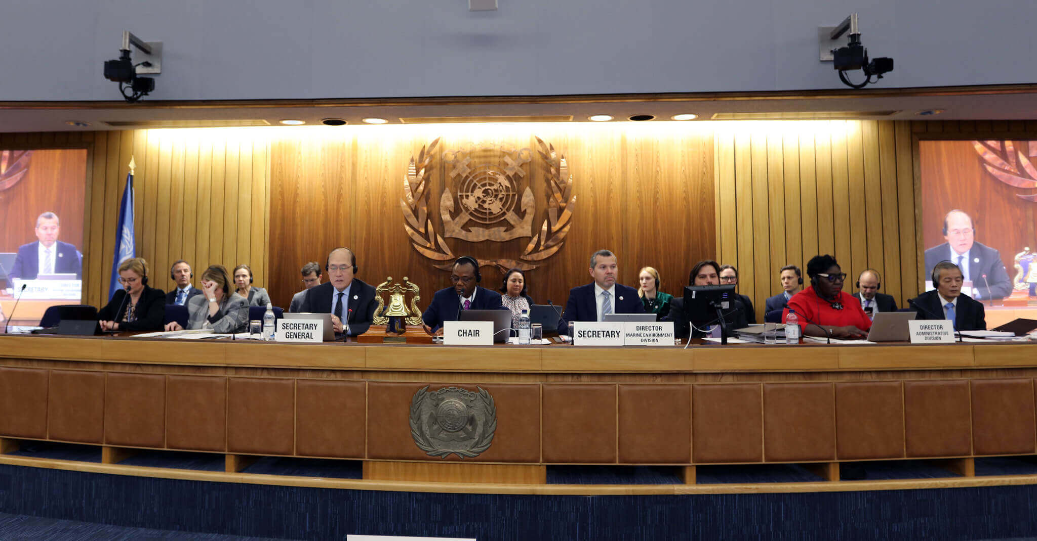 A panelist of leaders representing the International Maritime Organization sits behind a long wooden table in the IMO headquarters’s auditorium during the 79th Session of the Marine Environment Protection Committee. A large wood-carved IMO logo is seen at the center, behind the speakers.