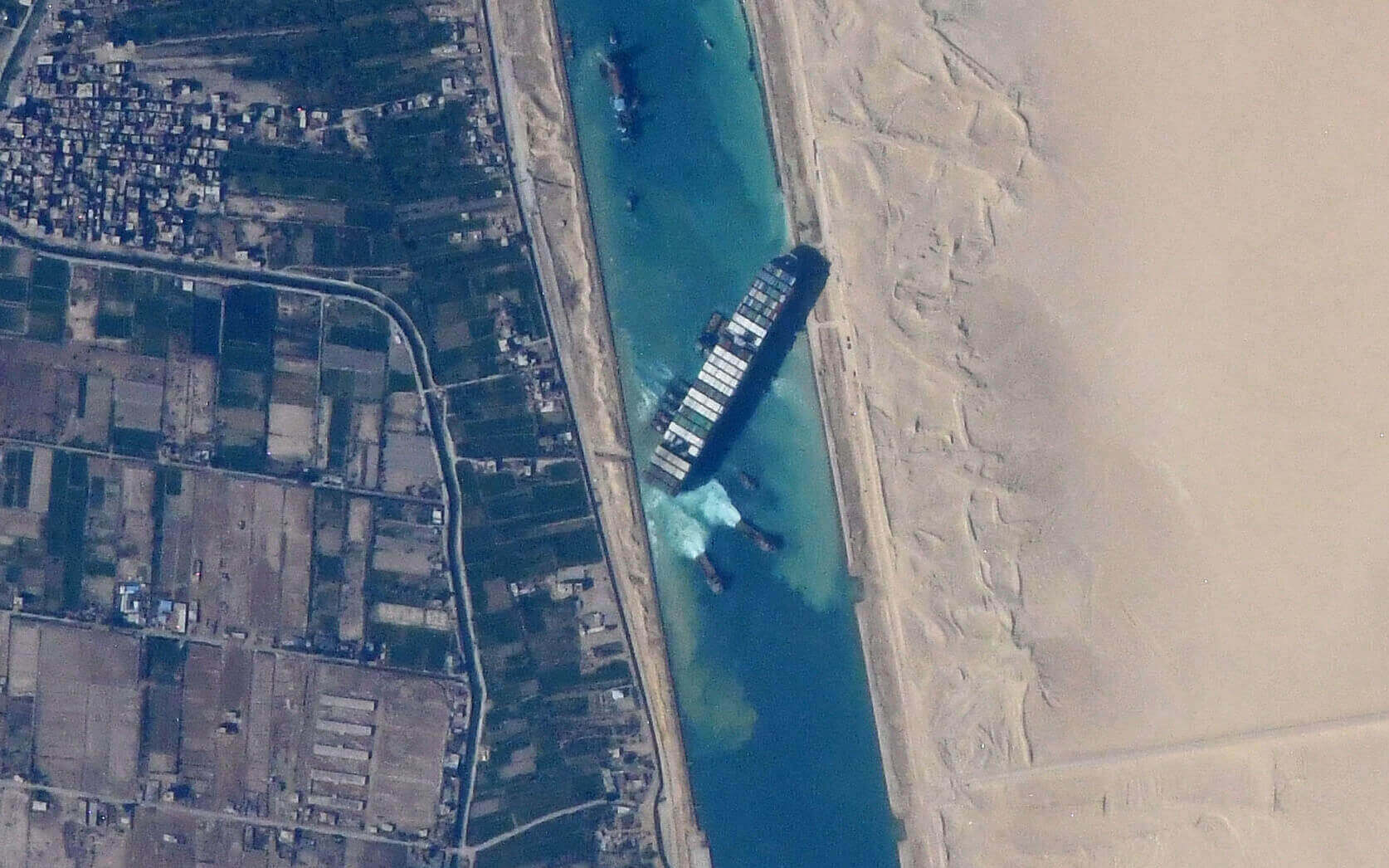 A satellite photo of the container ship Ever Given obstructing the green-hued Suez Canal. To the left of the canal are green agriculture fields. To the right are tan-colored sand dunes.