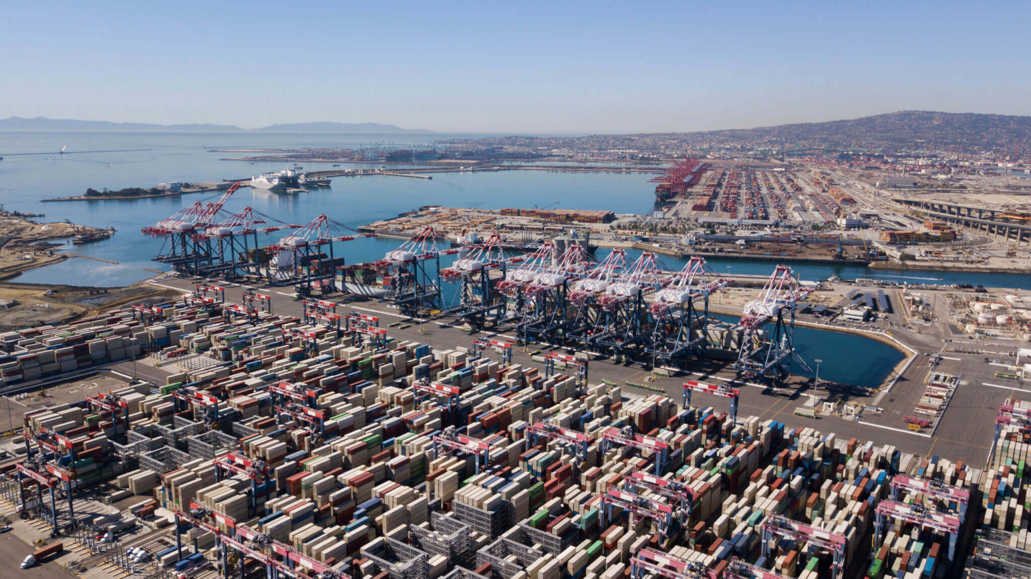 An aerial view of the Long Beach and Los Angeles ports filled with shipping containers. The Pacific Ocean and mountains are on the horizon.