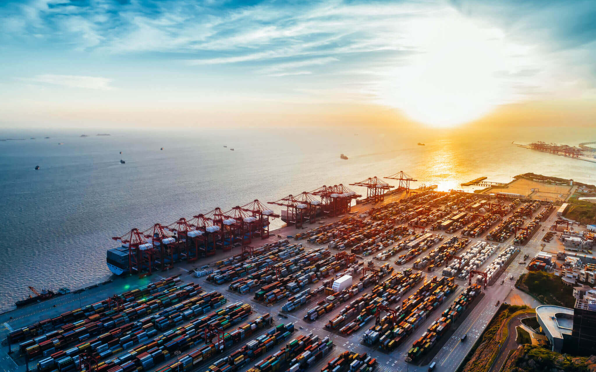 An aerial view of the Port of Shanghai. Visible are shipping containers lined up at the port with the East China Sea on the left and a golden sunset on the horizon to the right. 